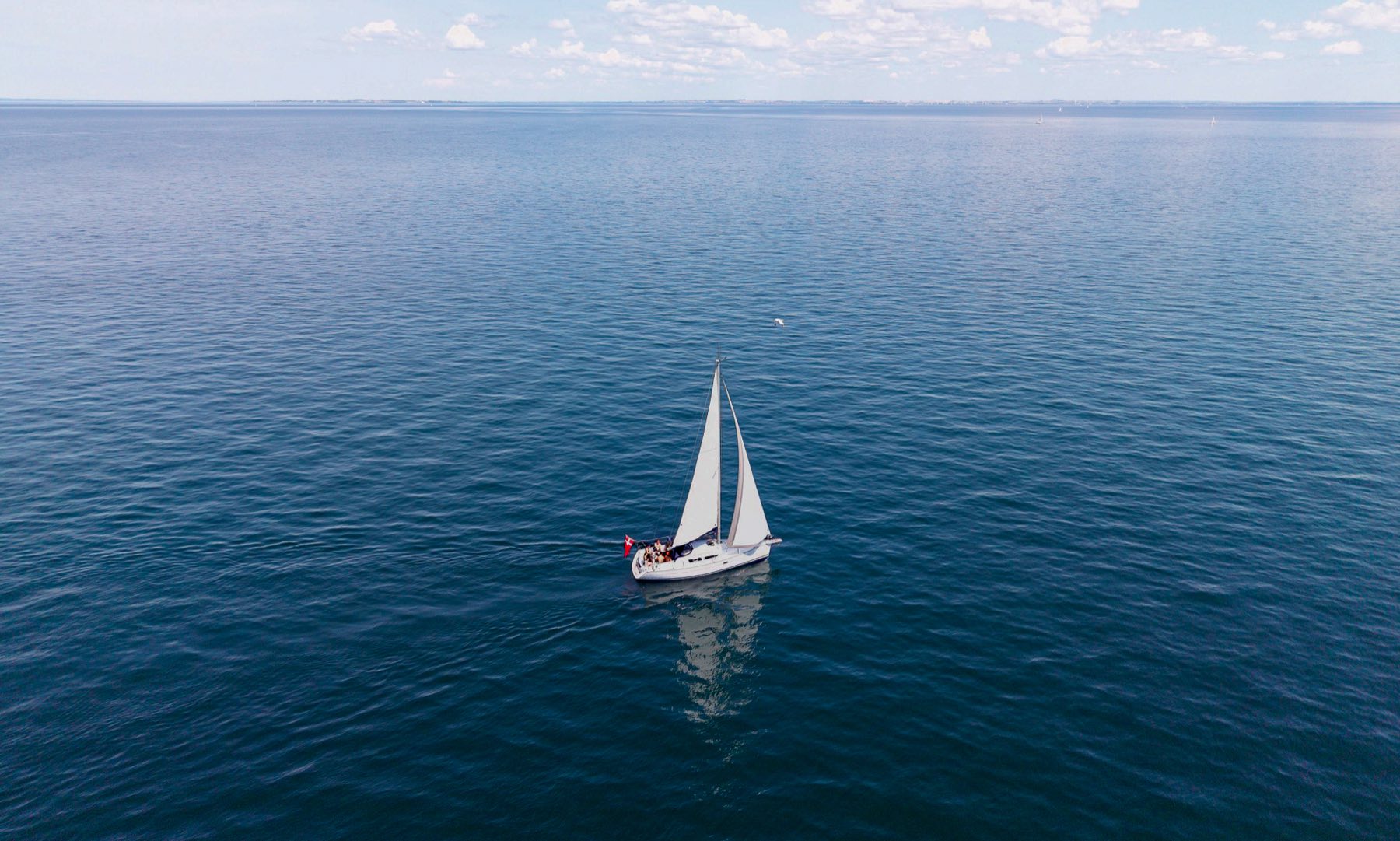 A sailboat under white sails on open blue water, Danish flag flying astern